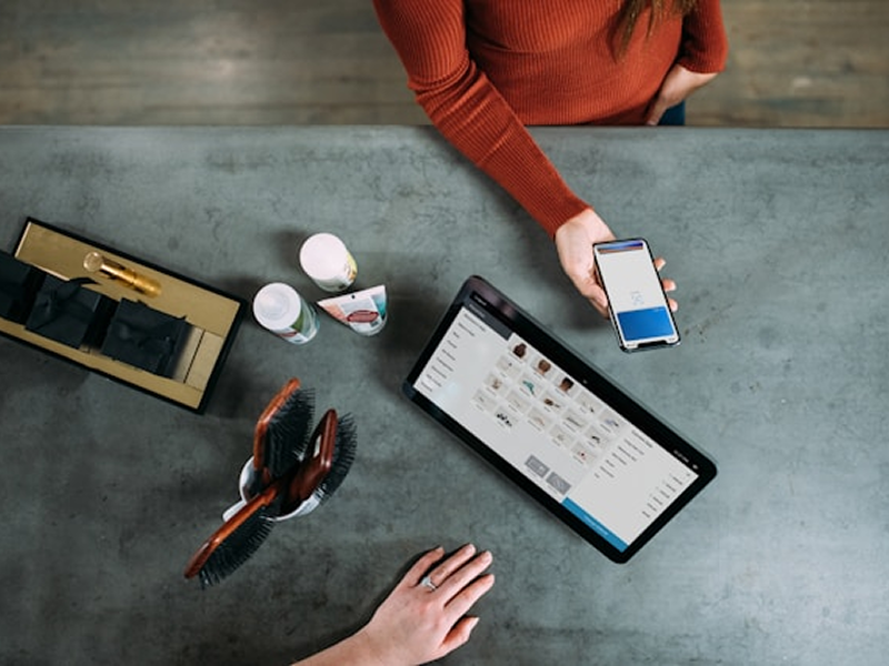 An overhead view of a retail checkout counter where a customer holds a smartphone to make a contactless payment, with a point-of-sale tablet and products on the counter.