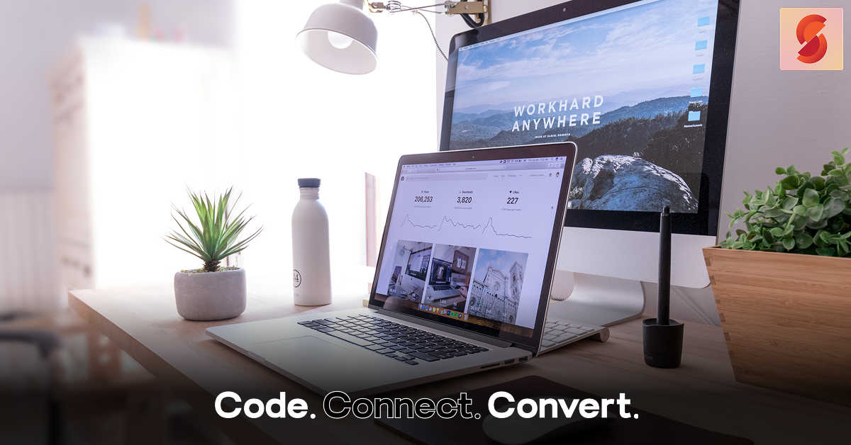 A modern and bright desk setup featuring a laptop displaying a data analytics dashboard, a large monitor with a "Work Hard Anywhere" wallpaper, and several potted plants.