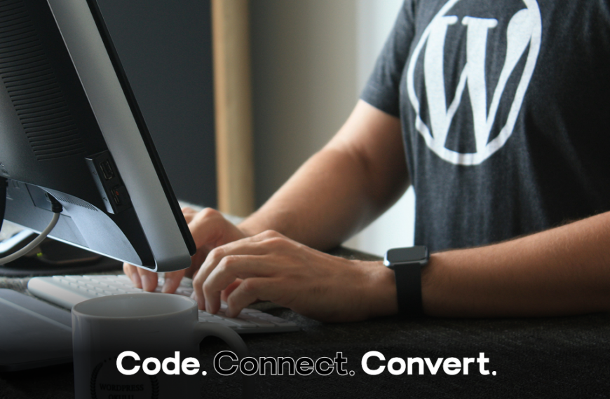 A person wearing a black t-shirt with the WordPress logo sits at a desk, typing on a white keyboard in front of a large monitor.