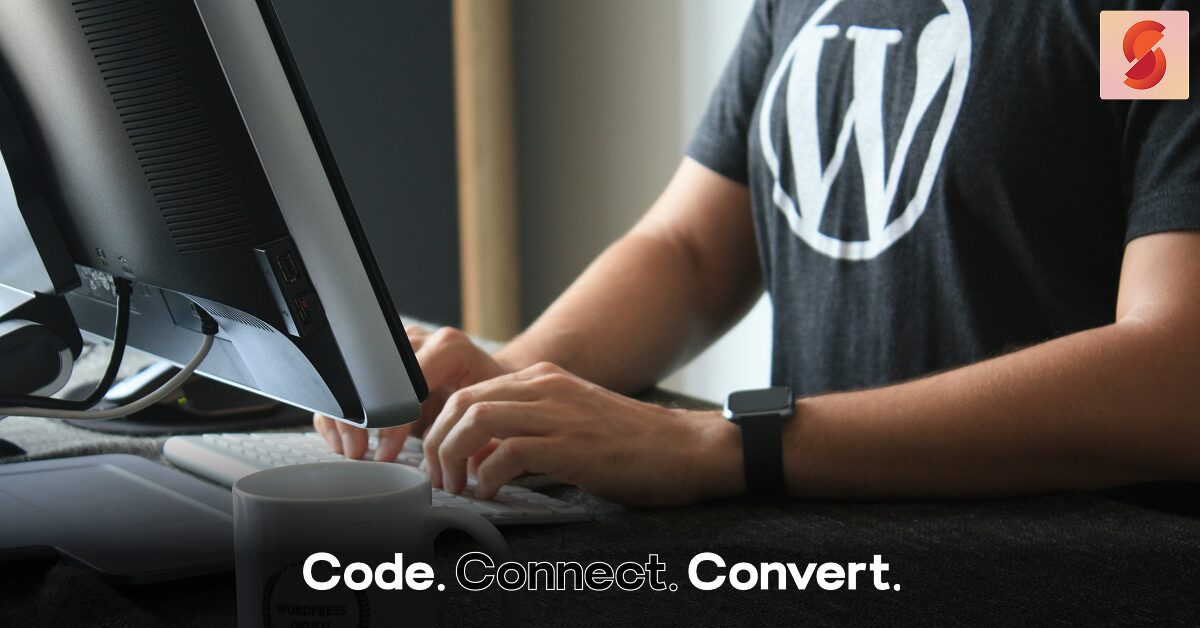 A person wearing a black t-shirt with the WordPress logo sits at a desk, typing on a white keyboard in front of a large monitor.