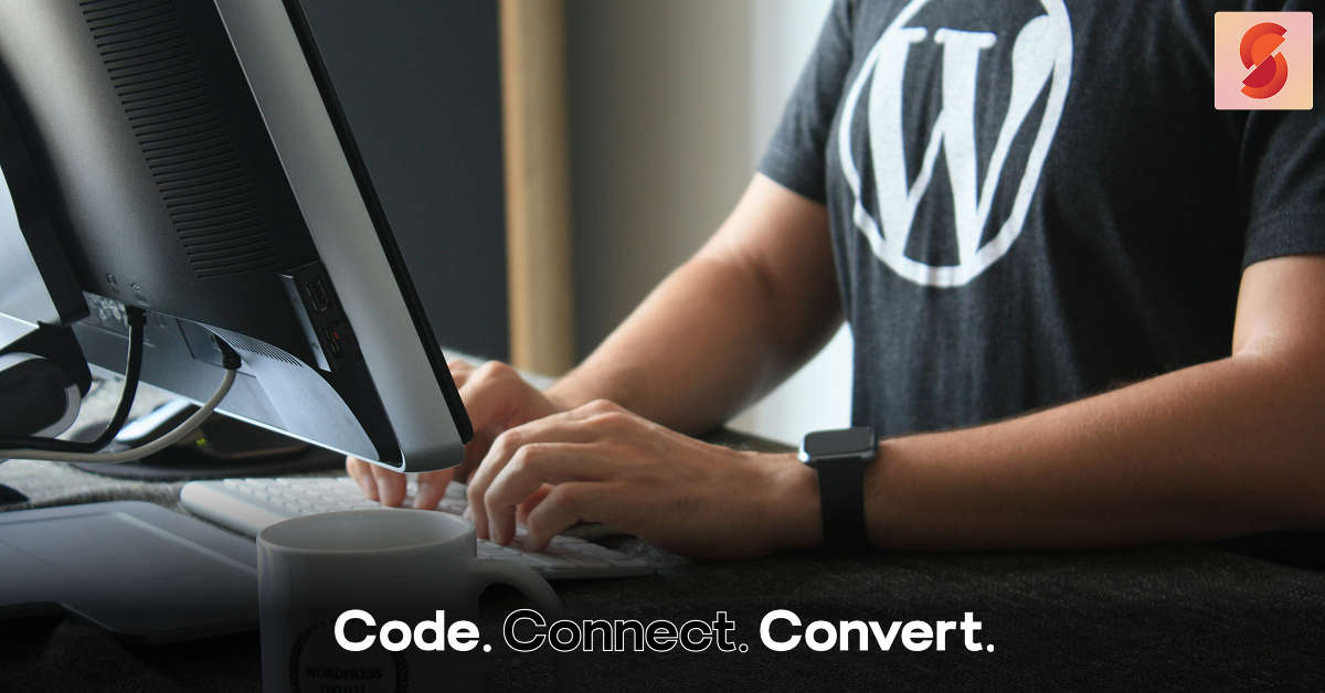 A person wearing a black t-shirt with the WordPress logo sits at a desk, typing on a white keyboard in front of a large monitor.