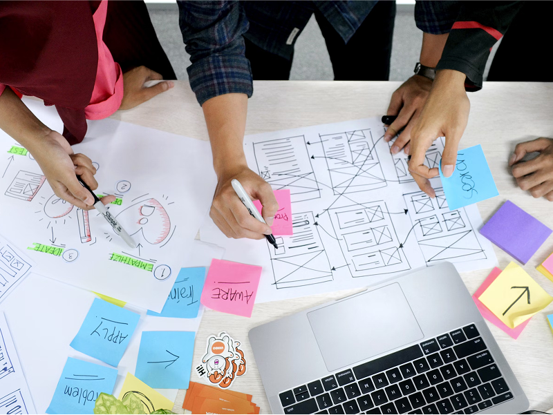 An overhead shot of a team's hands collaborating over hand-drawn website wireframes and diagrams, using markers and colorful sticky notes to map out ideas.