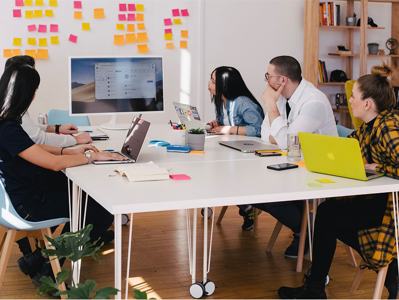 A diverse team of colleagues sits around a large white table in a bright meeting room, attentively looking at a large monitor, with a wall covered in colorful sticky notes behind them.