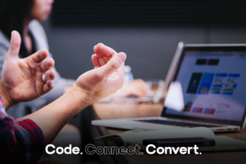 A person in a plaid shirt gestures with their hands while sitting at a wooden desk, with an open laptop and notebook visible in the background, suggesting a collaborative work meeting.