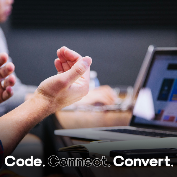 A person in a plaid shirt gestures with their hands while sitting at a wooden desk, with an open laptop and notebook visible in the background, suggesting a collaborative work meeting.