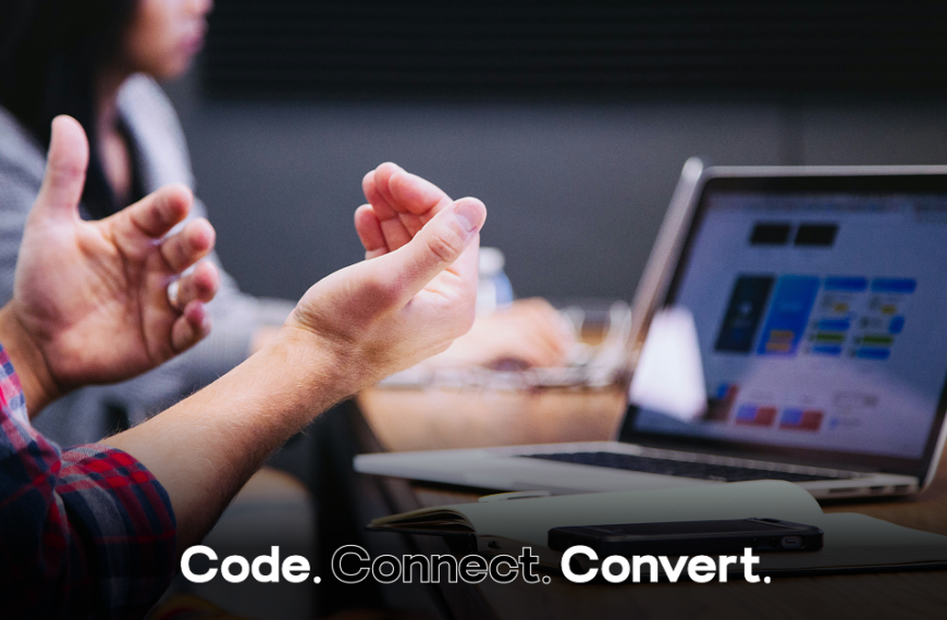 A person in a plaid shirt gestures with their hands while sitting at a wooden desk, with an open laptop and notebook visible in the background, suggesting a collaborative work meeting.