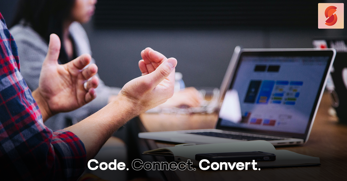 A person in a plaid shirt gestures with their hands while sitting at a wooden desk, with an open laptop and notebook visible in the background, suggesting a collaborative work meeting.