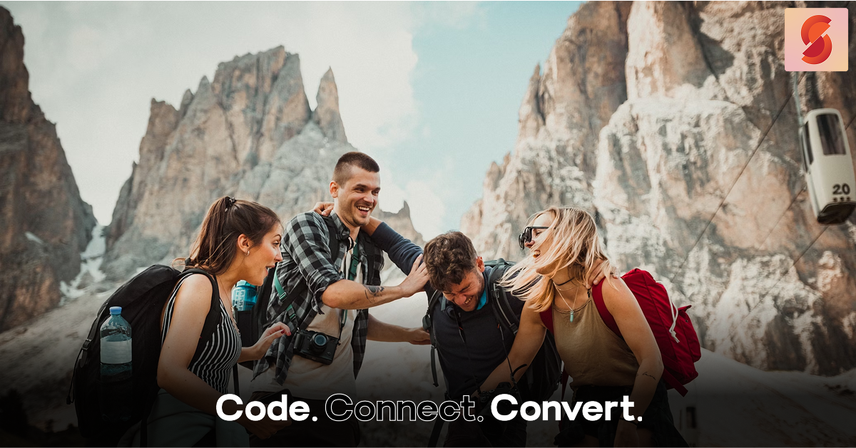 A group of four happy friends laugh together while hiking in a dramatic mountain setting, with a cable car visible in the background.