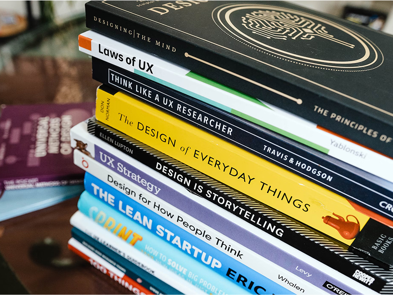 A tall stack of books focused on design and user experience, with titles including "The Design of Everyday Things," "Laws of UX," and "The Lean Startup."