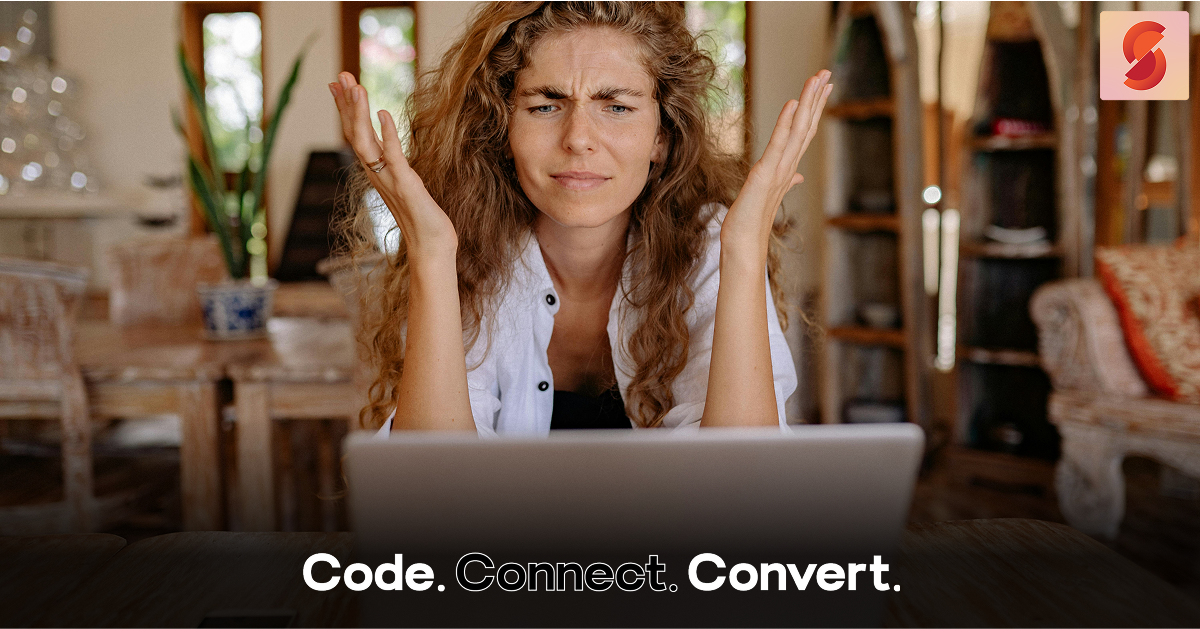 A woman with a confused and frustrated expression looks up from her laptop, holding her hands up in exasperation.