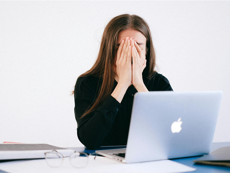 A woman in a black shirt sits at her desk in front of a laptop, covering her face with her hands in a gesture of stress or frustration.