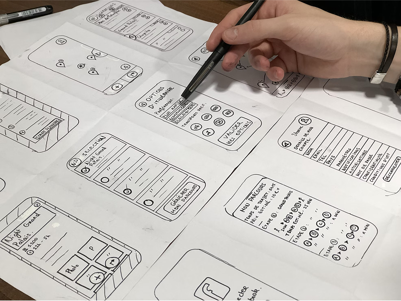 A designer's hand uses a pen to point at details on several hand-drawn wireframe sketches of a mobile application, which are laid out on a desk.