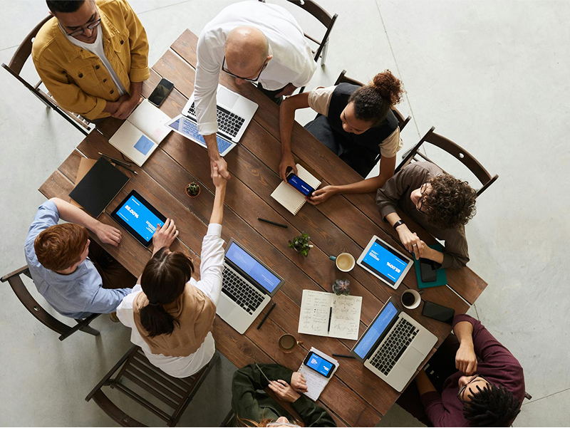 An overhead, bird's-eye view of a diverse business team sitting at a wooden table, with two members shaking hands in the center to signify a deal.