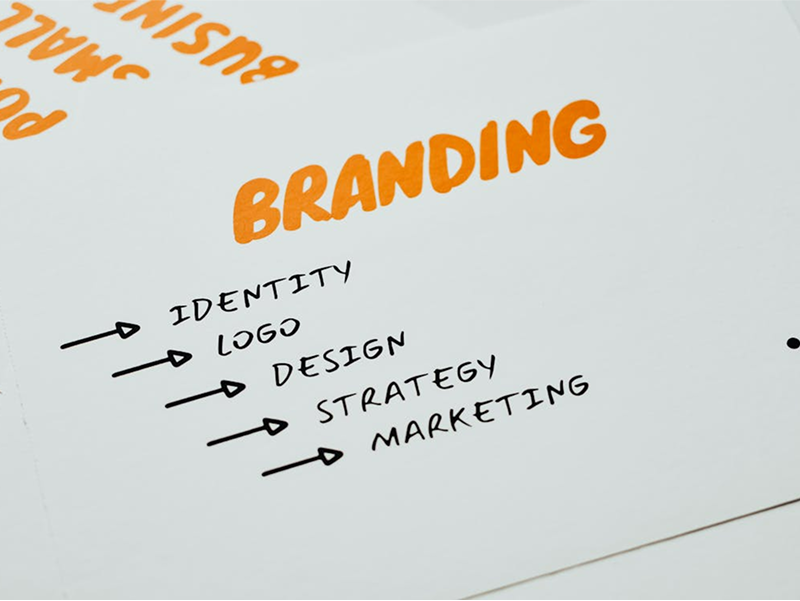 A close-up of a handwritten note with the word "BRANDING" in orange, followed by a list of key components: Identity, Logo, Design, Strategy, and Marketing.