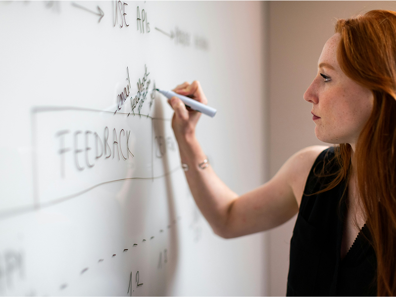 A focused woman with long red hair stands in profile, writing on a whiteboard with a marker, mapping out a process that includes the word "FEEDBACK.