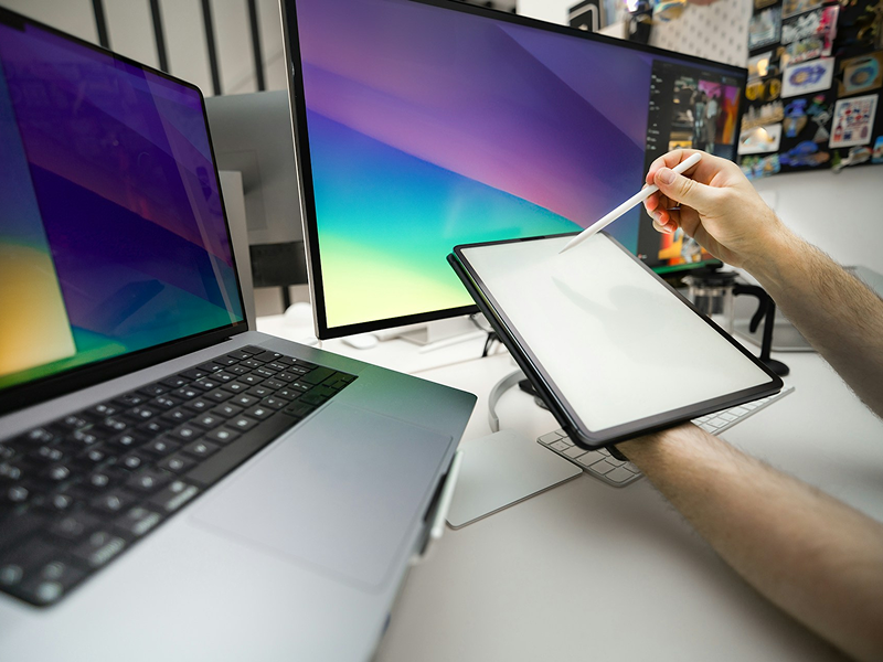 A close-up of a designer's desk with a laptop and large monitor, where a hand holds a stylus poised over a blank digital tablet.