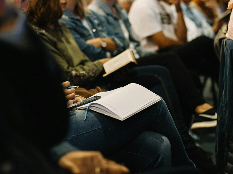 A close-up of a row of people sitting in an audience, with the focus on a person holding an open, blank notebook and a pen on their lap.