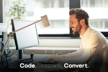 A smiling, bearded man in a white shirt sits at his desk in a modern office, happily looking at his computer screen while working.