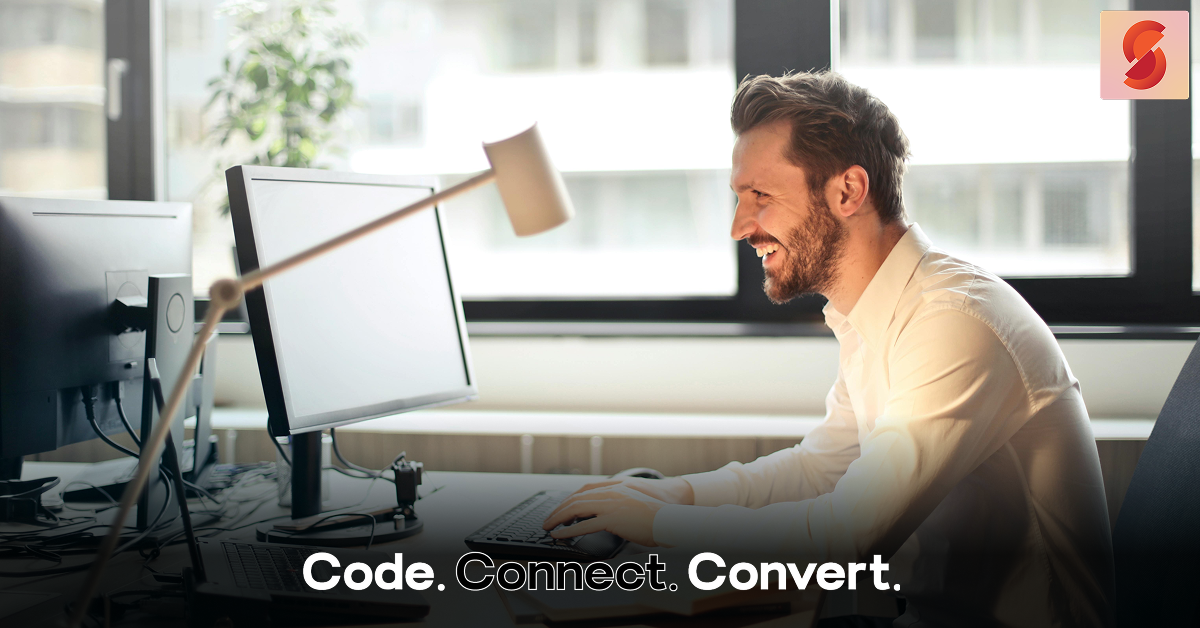 A smiling, bearded man in a white shirt sits at his desk in a modern office, happily looking at his computer screen while working.