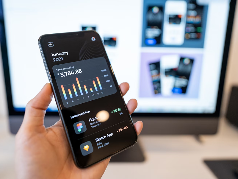 A hand holds a smartphone displaying a financial tracking app in dark mode, showing a monthly spending graph and recent transactions. A large computer monitor with design software is in the background.