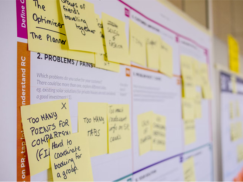  A close-up of a whiteboard from a workshop, covered in yellow sticky notes that identify customer "problems and pains," such as "Too many points for comparison."