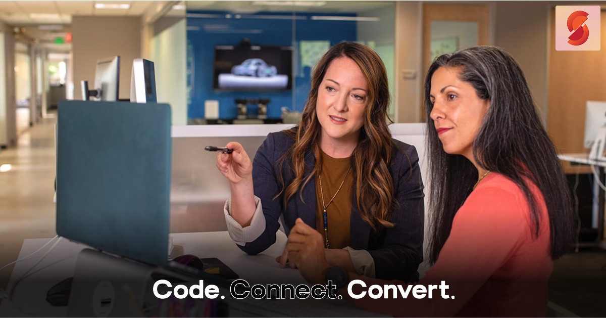 Two professional women collaborate in an office, with one pointing her pen at a computer screen to explain something while her colleague listens attentively.