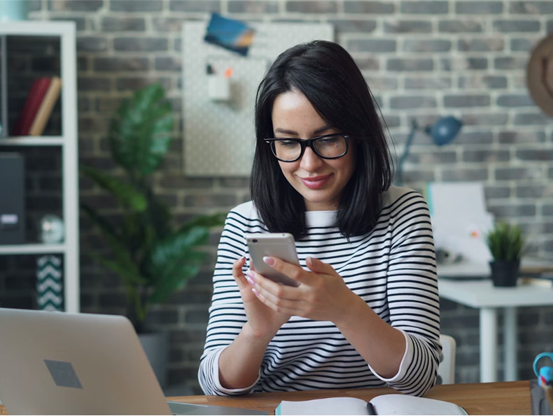 A smiling woman with dark hair and glasses sits at her office desk, looking down and engaging with her smartphone.