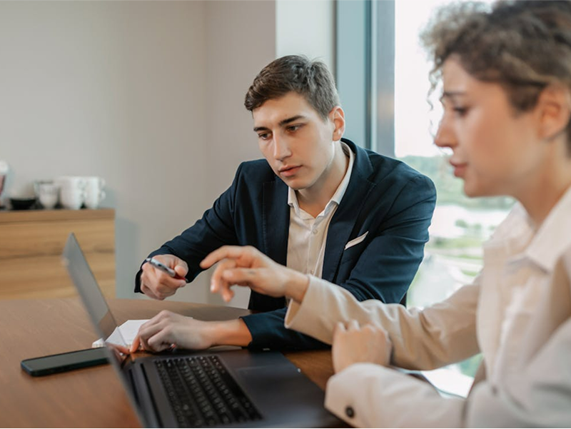  A man and a woman in business attire sit together at a desk, intensely focused on a laptop screen as the woman points out a detail to her colleague.