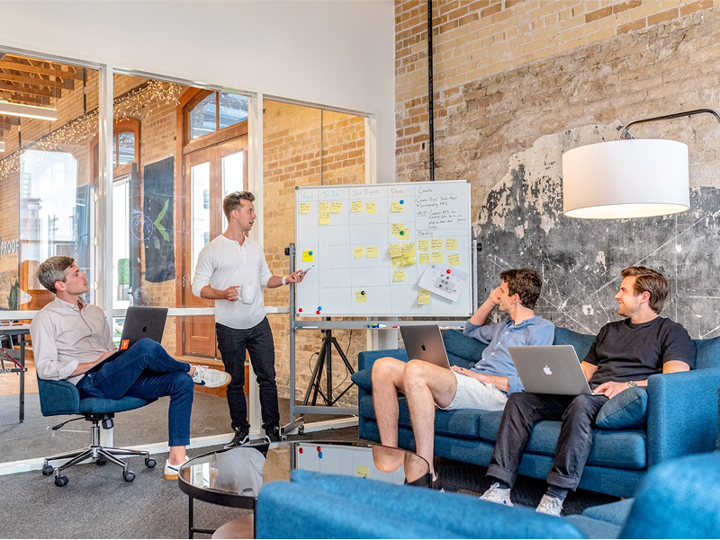Four casually dressed men having a team meeting in a modern office. One man stands and presents at a whiteboard covered with sticky notes, while the other three listen with laptops from a couch and chair.