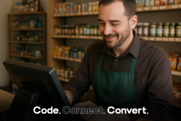 A smiling male shopkeeper wearing a green apron uses a point-of-sale computer terminal, with shelves of products visible behind him.