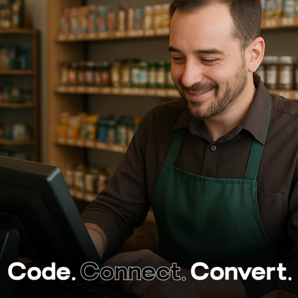 A smiling male shopkeeper wearing a green apron uses a point-of-sale computer terminal, with shelves of products visible behind him.