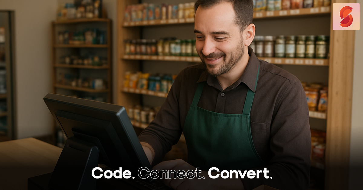 A smiling male shopkeeper wearing a green apron uses a point-of-sale computer terminal, with shelves of products visible behind him.