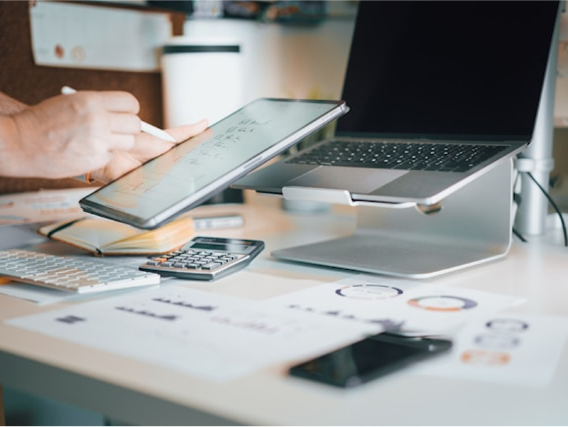 A person at a busy desk uses a stylus on a tablet, with a laptop on a stand, a calculator, and papers with charts spread out, indicating a multitasking work environment.