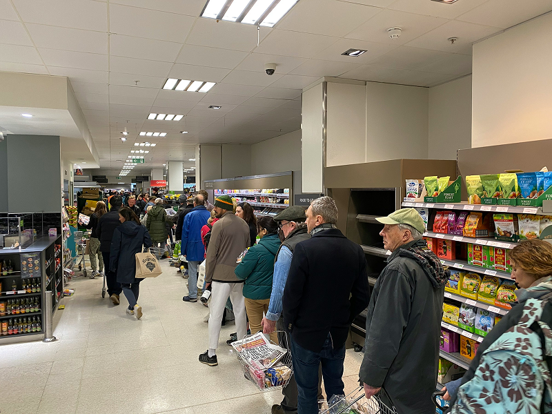 A long queue of shoppers with their baskets waiting in line inside a brightly lit supermarket aisle.