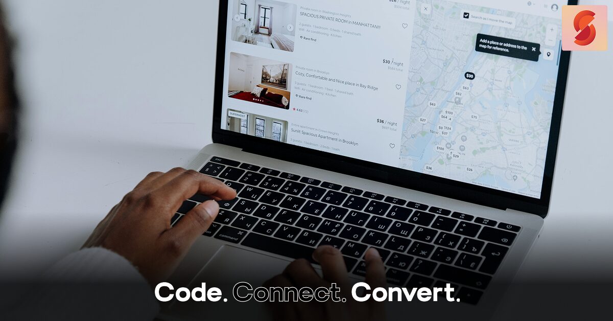 An over-the-shoulder view of a person's hands on a laptop keyboard, browsing a travel rental website that shows a list of properties and a city map.