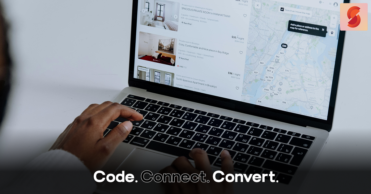 An over-the-shoulder view of a person's hands on a laptop keyboard, browsing a travel rental website that shows a list of properties and a city map.