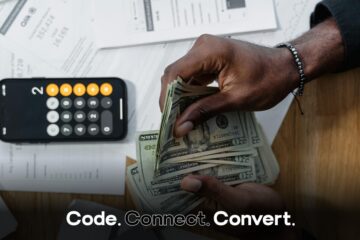 An overhead view of a person's hands counting a fan of US dollar bills over a desk scattered with financial documents and a smartphone showing a calculator.