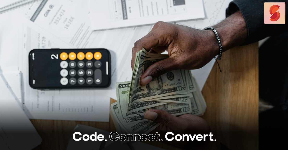 An overhead view of a person's hands counting a fan of US dollar bills over a desk scattered with financial documents and a smartphone showing a calculator.